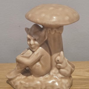 May include: A monochromatic beige ceramic figurine featuring a seated elf-like creature resting under a large mushroom. A small rabbit sits beside the mushroom stem on a textured base. The entire piece has a smooth, glossy finish and uniform colour.
