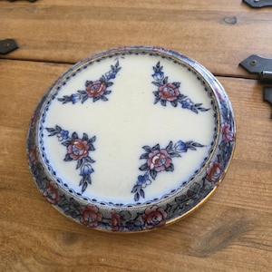 Può includere: Un piatto rotondo in ceramica con un centro color crema decorato con quattro gruppi floreali nelle tonalità del rosa, blu e grigio scuro. Il bordo esterno è ornato da un motivo floreale continuo e da un sottile bordo metallico dorato.