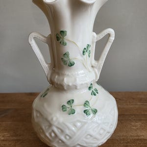 Könnte beinhalten: Eine cremefarbene Keramikvase mit gewelltem Rand, zwei seitlichen Henkeln und einem strukturierten Gittermuster am unteren Körper. Die Oberfläche ist mit handgemalten grünen Kleeblattmotiven verziert, die über den Hals und den bauchigen Fuß verteilt sind.