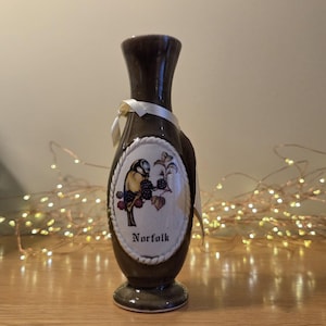 May include: A dark brown ceramic vase with a narrow neck and a flared base. An oval white plaque is centred on the front, featuring a colourful illustration of a bird perched on a blackberry branch with the word Norfolk printed below it.
