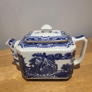 May include: A square ceramic teapot featuring a blue and white willow pattern with a landscape of trees, mountains, and buildings. The teapot has a matching lid with a small knob, a curved handle, and a short spout, all finished with gold trim.