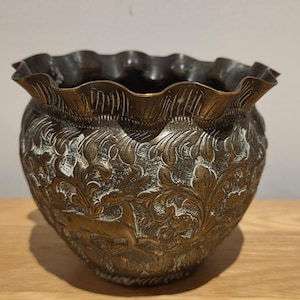 May include: A round brass bowl with a ruffled, scalloped rim. The exterior surface features an intricate, embossed pattern of stylized floral motifs and leaping deer figures, with a dark, oxidized finish highlighting the recessed details.