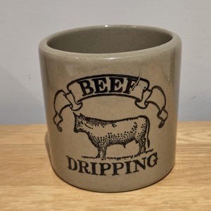 May include: A cylindrical beige ceramic crock with a speckled finish. The front features a black graphic illustration of a cow centred below a banner containing the word BEEF, with the word DRIPPING printed in black capital letters below the cow.