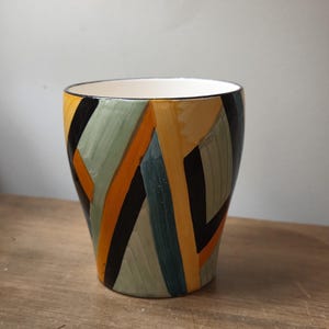May include: A ceramic plant pot with a geometric design. The pot has a white interior and a black rim. The exterior is decorated with vertical stripes in black, orange, green, and blue. The pot sits on a wooden surface.