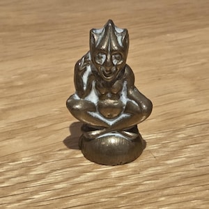 Op de afbeelding: Een klein, metalen beeldje in bronskleur dat een zittend wezen met puntige oren en een gehoornde kop voorstelt. Het figuurtje zit met gekruiste benen en één hand bij het gezicht, en heeft een verweerde afwerking met witte accenten.