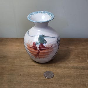 Puede incluir: Un pequeño jarrón de cerámica blanca con cuerpo redondo y cuello estrecho. Decorado con pinceladas abstractas en naranja, rojo y turquesa. El borde está pintado de azul claro. Una moneda está al lado del jarrón. El jarrón mide unos 10 cm de alto.