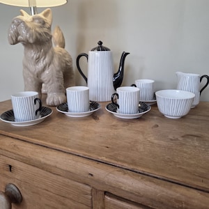 May include: A ceramic tea set featuring a white teapot, creamer, and four cups with saucers, all decorated with thin vertical black stripes and black handles. A beige ceramic dog figurine stands behind the tea set on the wooden surface.