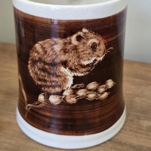 Peut inclure: Mug en céramique avec un motif marron et blanc. La tasse présente une illustration détaillée d'une souris brune mangeant des graines. Le fond est de couleur marron foncé, et le bord et la base sont blancs.