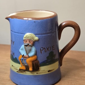 May include: A blue ceramic pitcher with a brown handle and rim. The pitcher features a relief of a seated gnome with a white beard and a green hat. The word "Pixie" is written on the side.
