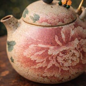 May include: A ceramic teapot with a floral design in shades of pink and cream. The teapot has a dark blue knob on the lid and a gold-colored handle. The design includes green leaves and a decorative charm.