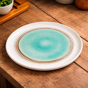 Puede incluir: Un plato de cerámica blanca con un centro turquesa y círculos concéntricos marrones. El plato tiene un borde ligeramente elevado y está sobre una superficie de madera. El plato es parte de un juego de vajilla.