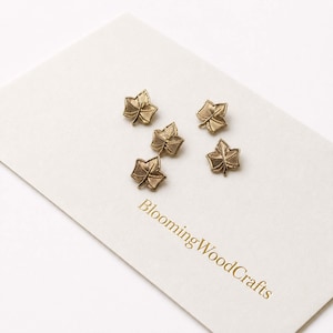 May include: Four small, metallic gold-toned leaf-shaped charms arranged on a white card. The card features the text BloomingWoodCrafts printed in gold lettering. Each leaf charm displays detailed, embossed vein patterns across its surface.