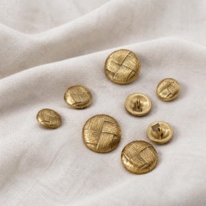 May include: A collection of round, metallic gold-toned buttons featuring a textured, woven-style pattern on the surface. Several buttons are displayed from the front, while others are flipped to show the metal shank attachment on the back.