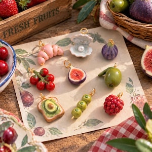 May include: A collection of miniature food charms including a pink croissant, an open oyster shell with a pearl, a purple fig, a cluster of red tomatoes, a sliced fig, a green olive, avocado toast, a skewer of three olives, and a red raspberry.