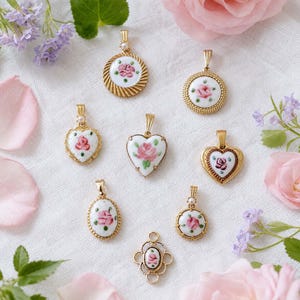 May include: A collection of seven gold-toned metal pendants featuring white ceramic centers with hand-painted pink rose motifs and green leaves. The pendants include circular, heart-shaped, and oval designs, each with a small bail for attachment.