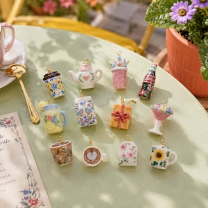 May include: A collection of twelve miniature ceramic charms featuring various designs including a teapot, a soda bottle, a gift box, a floral mug, a lemon jar, a milk carton with birds, a coffee cup with heart art, and a sunflower mug with gold loops.