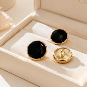 Può includere: Tre bottoni circolari con un inserto centrale nero liscio e lucido, circondato da un bordo in metallo color oro lucido. Un bottone è posizionato in modo da mostrare il retro metallico piatto con un piccolo occhiello centrale per il fissaggio.