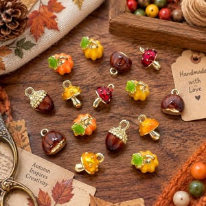 May include: A collection of small metal charms featuring autumn-themed designs including brown acorns, orange pumpkins with green stems, and red or yellow mushrooms with white spots, each equipped with a small loop for attachment to jewellery.