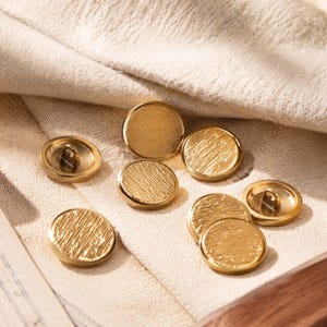 May include: A collection of seven round, gold-coloured metal buttons featuring a textured, brushed surface finish. Each button has a smooth, raised edge and a shank on the back for attachment, displaying a consistent metallic lustre and circular shape.
