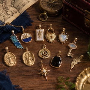 Puede incluir: Una colección de colgantes de metal dorado con varios diseños, incluyendo un ala azul, un sol, un corazón con espada, un candado, un corazón con ópalo, una llave, una serpiente, una piedra negra, una estrella y dos figuras ovaladas.
