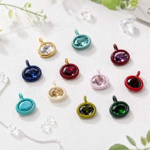 May include: A collection of twelve circular jewellery charms, each featuring a faceted gemstone set within a coloured metal frame. The charms display a variety of vibrant hues including blue, red, green, yellow, pink, and clear, each with a small loop.