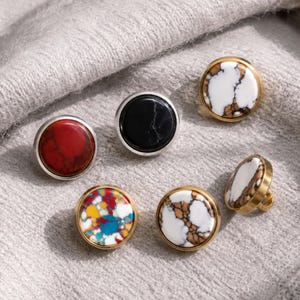 May include: A collection of six round buttons featuring various stone-like patterns. The set includes one red, one black, one multicoloured, and three white buttons with brown veining, all set in metallic circular frames with visible back shanks.