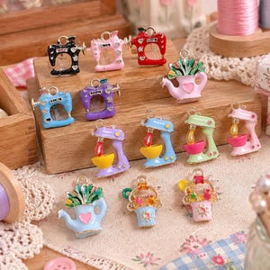 May include: A collection of metal charms featuring miniature sewing machines, stand mixers, and watering cans with colorful enamel finishes. The charms include various designs like pink, blue, red, and black sewing machines, pastel mixers, and watering cans with floral accents.