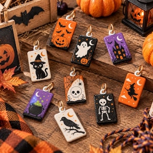 May include: A collection of nine rectangular Halloween-themed charms featuring illustrations of a black cat, a ghost, a haunted house, a cauldron, a skull, a skeleton, a witch, a raven, and a pumpkin, each with a gold-toned metal jump ring.