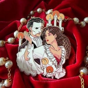 May include: Enamel pin depicting characters from the Phantom of the Opera, with the male figure in a mask and tuxedo. The female character wears a white dress. The pin includes candles and is set against a red velvet backdrop.