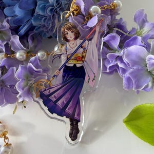 May include: A clear acrylic charm featuring a female anime character in a purple and white outfit, holding a staff. The charm is hanging from a gold-colored clasp. The background includes blue and purple flowers and faux pearls.