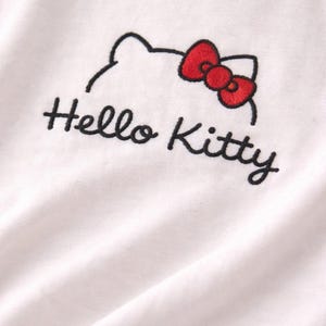 Puede incluir: Camiseta blanca con un dise&ntilde;o bordado de Hello Kitty. El dise&ntilde;o incluye un contorno negro de la cabeza de Hello Kitty y un lazo rojo. Las palabras "Hello Kitty" est&aacute;n bordadas en negro debajo.