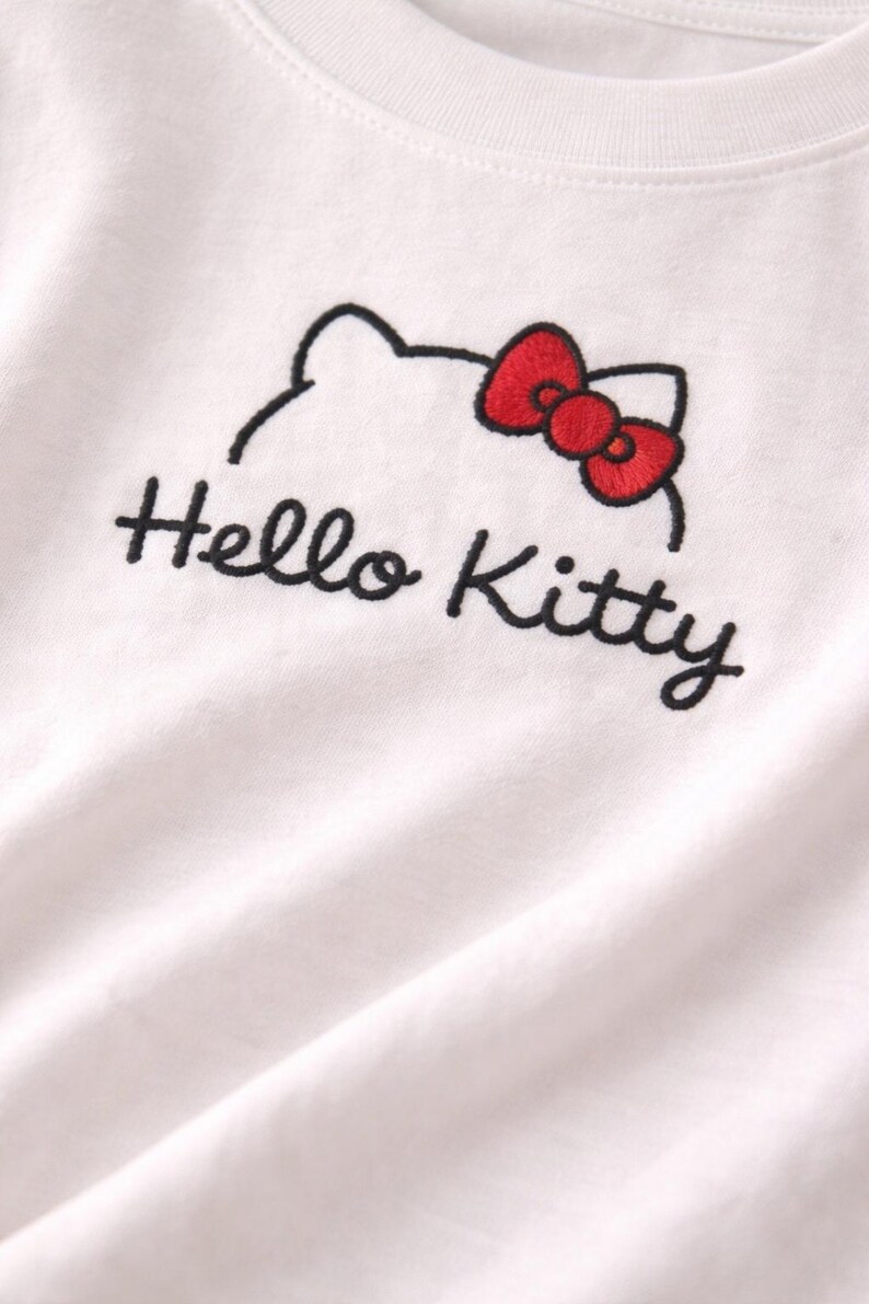 Puede incluir: Camiseta blanca con un dise&ntilde;o bordado de Hello Kitty. El dise&ntilde;o incluye un contorno negro de la cabeza de Hello Kitty y un lazo rojo. Las palabras "Hello Kitty" est&aacute;n bordadas en negro debajo.