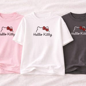 Puede incluir: Tres camisetas de manga corta en rosa, blanco y gris carb&oacute;n. Cada camiseta presenta un gr&aacute;fico de Hello Kitty con un lazo rojo y el texto "Hello Kitty" en escritura negra. Las camisetas est&aacute;n hechas de un material suave.