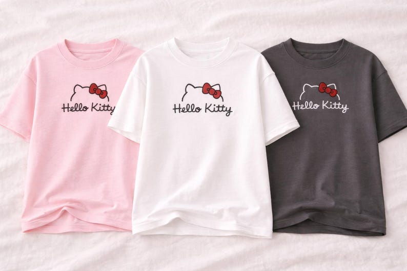 Puede incluir: Tres camisetas de manga corta en rosa, blanco y gris carb&oacute;n. Cada camiseta presenta un gr&aacute;fico de Hello Kitty con un lazo rojo y el texto "Hello Kitty" en escritura negra. Las camisetas est&aacute;n hechas de un material suave.