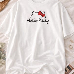 Puede incluir: Camiseta blanca con el personaje de Hello Kitty. El dise&ntilde;o incluye un contorno negro de la cara de Hello Kitty con un lazo rojo y las palabras "Hello Kitty" en escritura negra. La camiseta es de material suave.