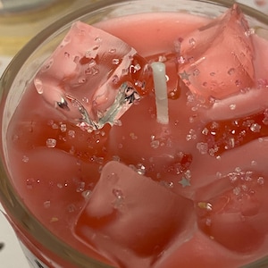 May include: A close-up of a candle in a clear glass jar. The candle wax is a light pink color, with clear, ice-like shapes and small, sparkly embellishments. A white wick is centered in the wax.