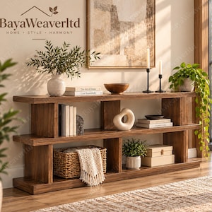 May include: A long, low-profile wooden console table with two open shelves. The unit features a dark wood grain finish and a tiered design. It is styled with books, a woven basket with a cream throw, potted plants, candles, and decorative objects.