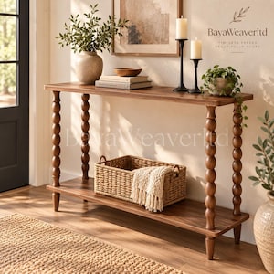 May include: A rectangular wooden console table with four turned spindle legs and a lower shelf. The table holds a ceramic vase with greenery, two stacked books, a wooden bowl, and two black candle holders. A woven basket with a cream throw sits below.