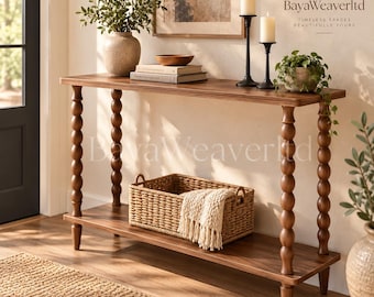 May include: A rectangular wooden console table with four turned spindle legs and a lower shelf. The table holds a ceramic vase with greenery, two stacked books, a wooden bowl, and two black candle holders. A woven basket with a cream throw sits below.