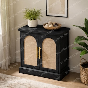 May include: A black wooden cabinet featuring two doors with arched natural rattan panels and vertical gold metal handles. The top surface holds a white ceramic pot with a green plant and a wooden bowl containing decorative spheres.