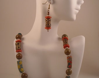 May include: A necklace and earrings set featuring brown, orange, and green ceramic beads. The necklace has a variety of bead shapes and sizes, including round, cylindrical, and patterned beads. The earrings are simple and feature a single cylindrical bead.