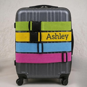 May include: A gray suitcase secured with four colorful luggage straps. The straps are green, yellow with the name "Ashley", blue, and pink. Each strap has a black buckle for closure.
