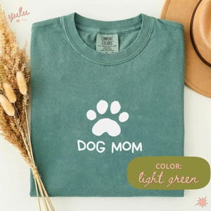 Può includere: Una t-shirt a maniche corte color verde chiaro con una grafica bianca di un'impronta di zampa di cane sopra la scritta "DOG MOM" in un carattere casual. La maglietta presenta un girocollo e un'etichetta del marchio Comfort Colors all'interno.