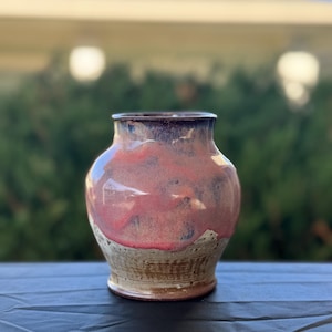 Può includere: Un vaso in ceramica fatto a mano con un corpo arrotondato e un bordo leggermente svasato. Il vaso presenta una tavolozza di colori rosa-rossastro, marrone e crema. Il bordo superiore è viola scuro. Adatto per fiori o come elemento decorativo.