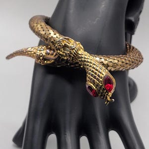 May include: A gold-toned metal snake bracelet featuring a textured, scale-like surface and two prominent red marquise-cut stones set as eyes. The serpent design wraps around the wrist, with the head and tail meeting at the front of the piece.