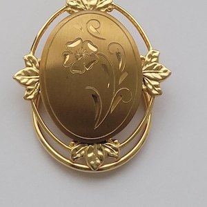 Può includere: Medaglione ovale dorato con un design floreale. Il medaglione presenta un fiore e uno stelo incisi, circondati da una cornice decorativa con accenti di foglie. Un gioiello classico.