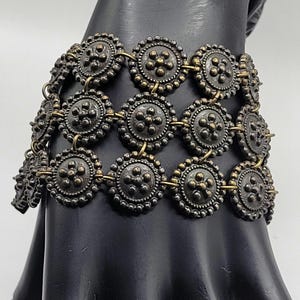 May include: A multi-strand bracelet composed of three rows of circular, dark metallic medallions. Each medallion features a central raised dot surrounded by five smaller dots, with a beaded edge, all connected by small brass-colored metal jump rings.