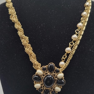 Puede incluir: Collar dorado con un colgante floral central con piedras negras y crema. El collar tiene una cadena trenzada en un lado y una cadena en capas con perlas de imitación en el otro, exhibido en un maniquí negro.