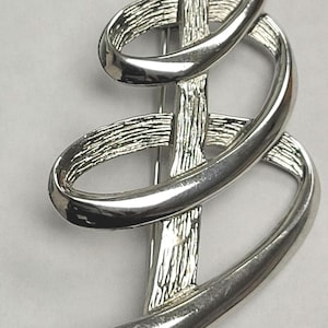 May include: Silver brooch featuring a textured central bar intersected by two smooth, looping curves. The abstract design of the brooch offers a contemporary aesthetic, ideal for enhancing various attire.