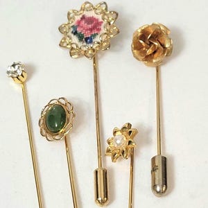 Könnte beinhalten: Sammlung von Vintage-Hutnadeln. Enthält Nadeln mit einem klaren Strassstein, einem grünen Stein, einem floralen Wandteppich, einer goldenen Rose und einer Perle. Die Nadeln haben goldfarbene Metallstiele und dekorative Köpfe. Ideal für Sammler.